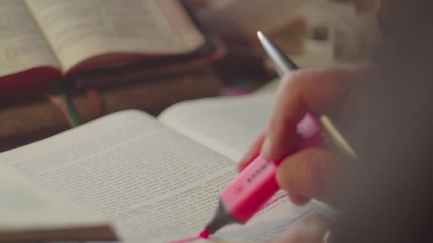 A closeup shot shows a persons hand using a pink highlighter to mark a line of text in an open book several other open books are visible in the background suggesting a study session