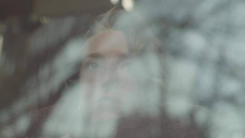 A young blonde woman is looking through a window with a pensive expression
