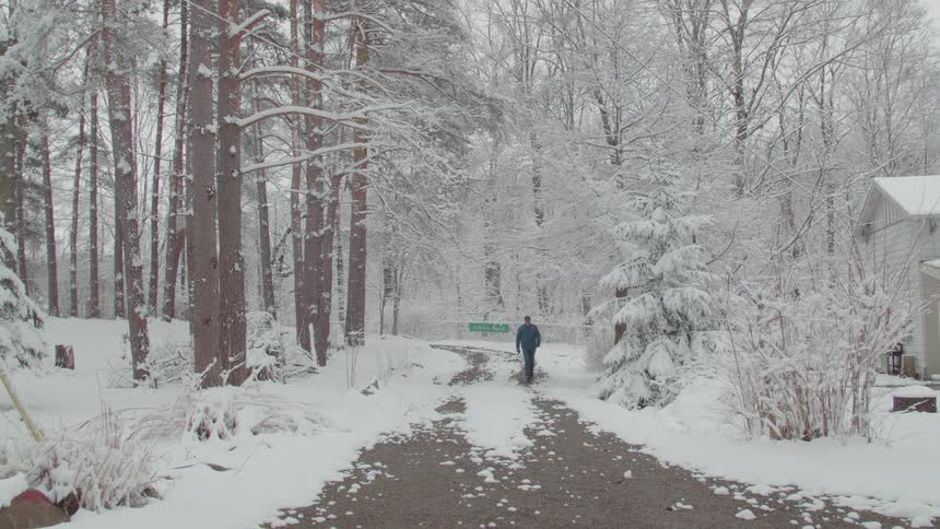 Man wearing a baseball cap and a blue jacket walking on a path surrounded by snow with a forest and a house to the sides outdoors during day time