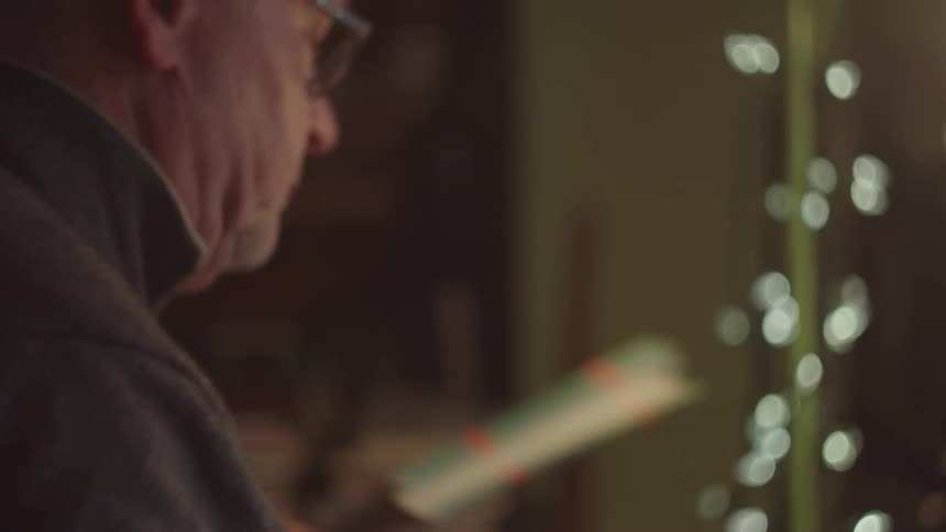 An older man wearing glasses looks down in concentration softly illuminated by warm indoor light