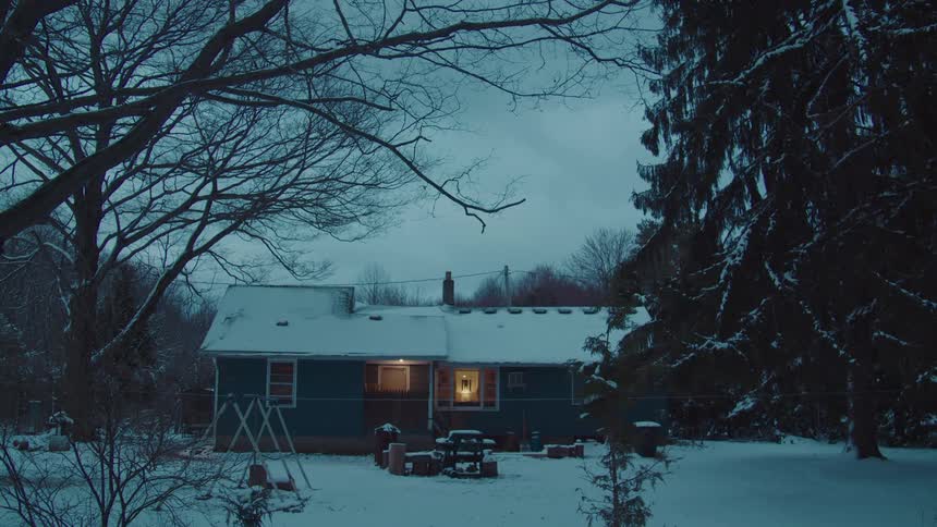 The video shows a singlestory house at dusk with its roof and surrounding grounds covered in snow a single line of light emanates from a window on the lower left of the house