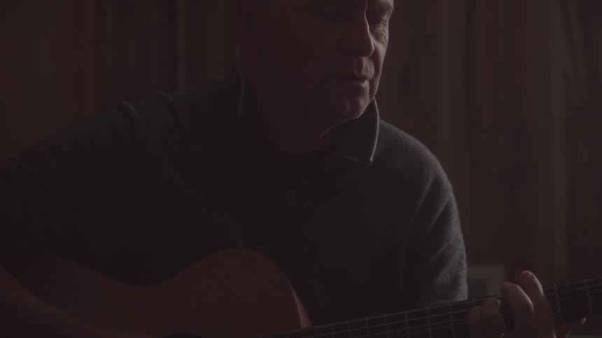 A closeup shot of a man playing an acoustic guitar indoors the man is wearing a gray sweater and has his head tilted down as he concentrates on the song