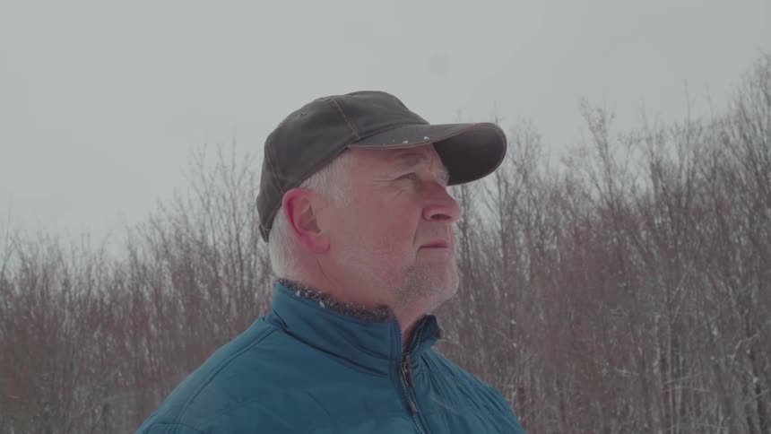 A shot from the chest up shows a man with a gray beard and white hair wearing a dark cap and a blue jacket he is standing in a field of dry tall grass under an overcast sky