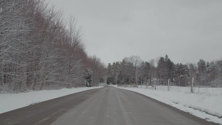 Paved road surrounded by a field covered in snow and leafless trees