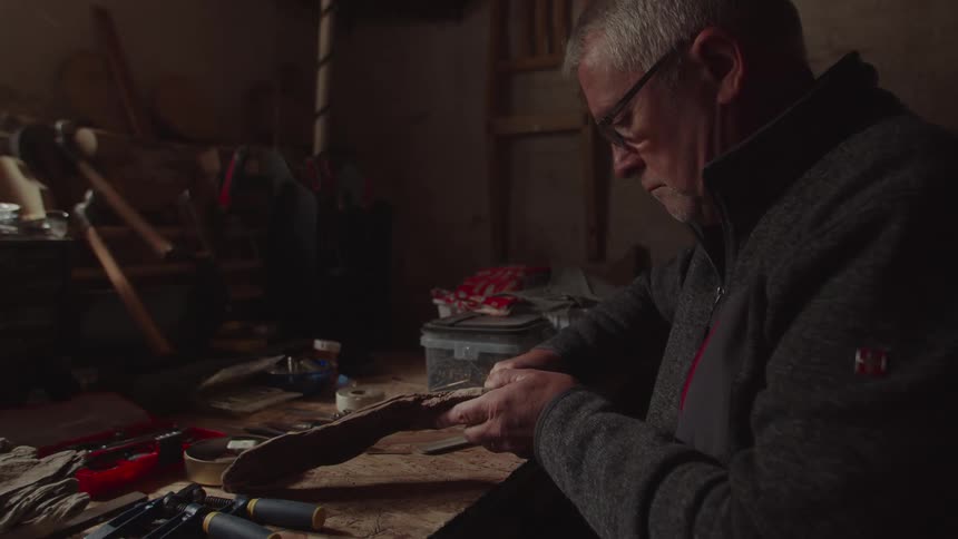 Elderly man wearing glasses and a jacket sitting down working on a piece of wood indoors