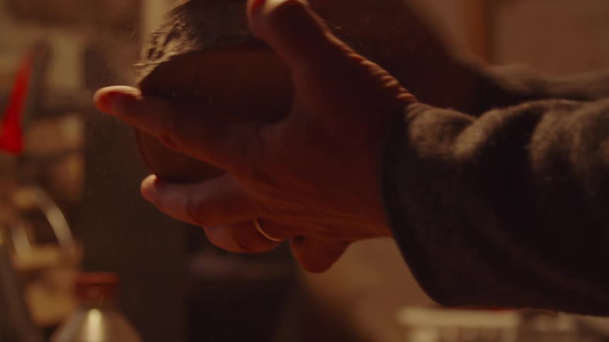 A persons hands are shown sanding a rough dark piece of wood in a dimly lit workshop