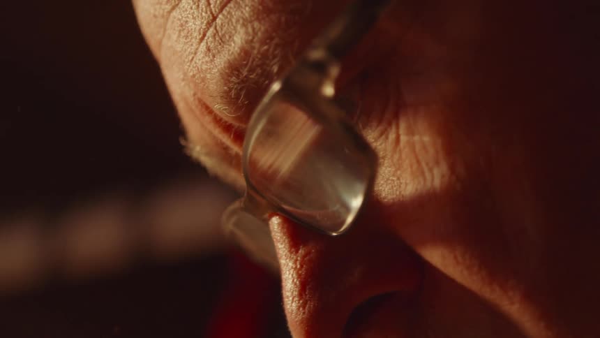 A macro shot shows a mans face in profile with a focus on his glasses and the wrinkles around his eyes a wisp of smoke drifts into the frame near his nose