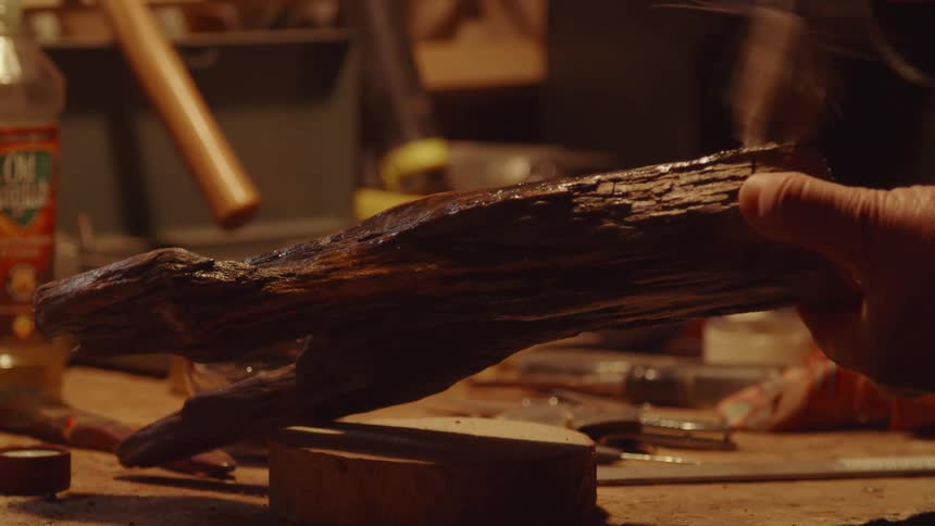 A close view shows a persons hands in a workshop carefully painting a piece of dark polished wood with a brush over a workbench filled with tools