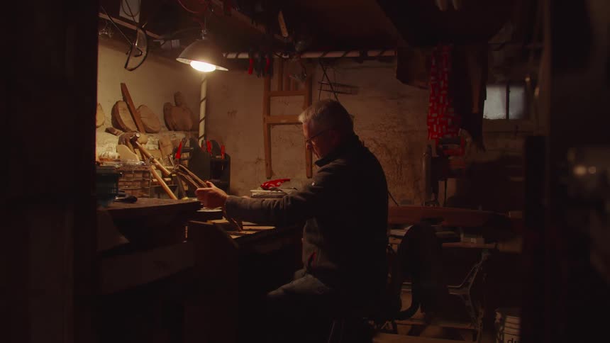 Elderly man sitting down on a chair polishing a piece of wood on a workshop indoors