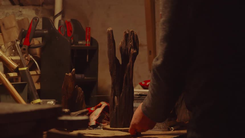 A persons hand reaches for a piece of wood on a workbench the workbench is cluttered with tools a plastic container of screws and other wooden pieces the scene takes place in a dimly lit workshop