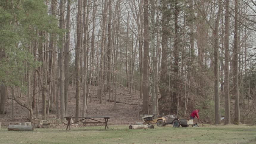 A person in a red jacket rakes leaves in a large yard with a small tractor and a picnic table all set against a backdrop of a dense forest