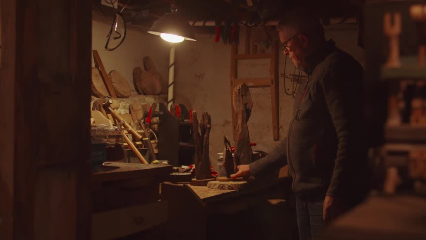 Elderly man wearing glasses standing up on a workshop analyzing a piece of wood on a stand indoors