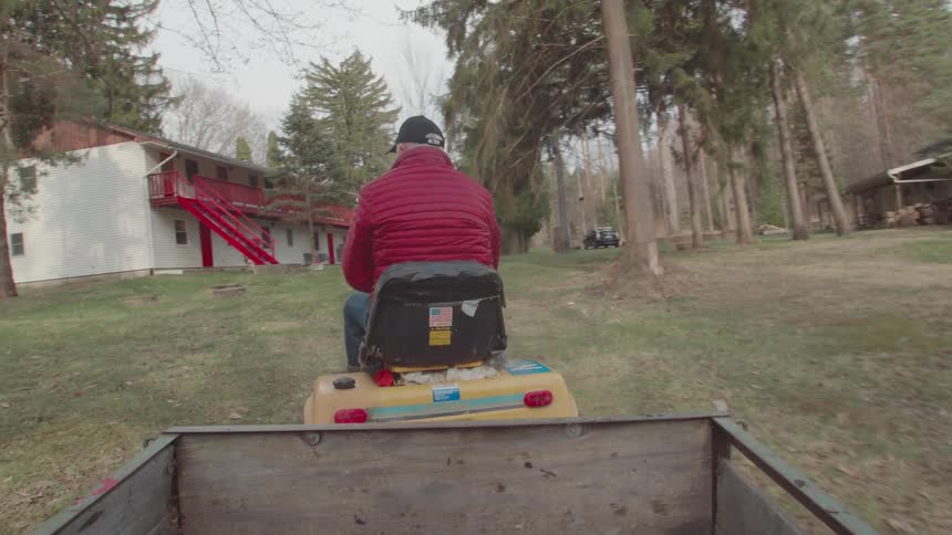 Man in a red jacket drives a lawn mower with a trailer in a wooded area