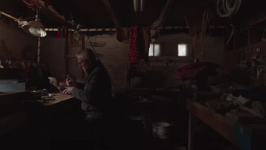 A person is seen from the side sitting at a workbench and working on a small object the room is dimly lit but a bright lamp hangs over the work area the workshop appears to be in disarray with various tools and materials scattered around