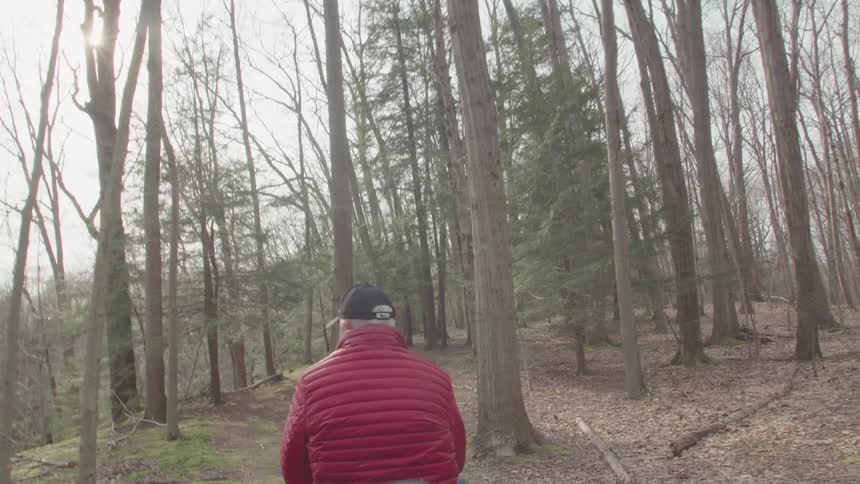 A shot from a drone follows a person wearing a red jacket and a black beanie as they walk through a forest the sun shines brightly through the trees creating lens flare