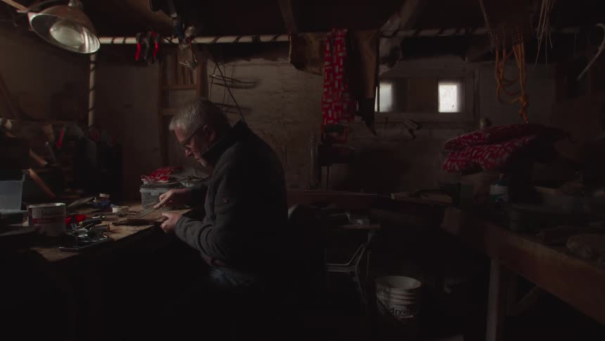 Elderly man wearing glasses and a jacket sitting down indoors working on a piece of wood