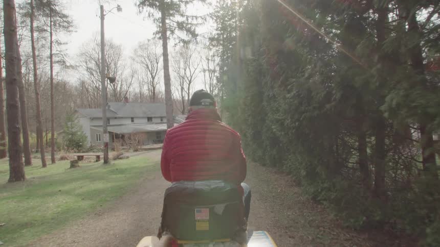 A wide firstperson shot shows a persons back as they ride on a small vehicle possibly a lawnmower down a path the path is lined by trees with a house visible in the background