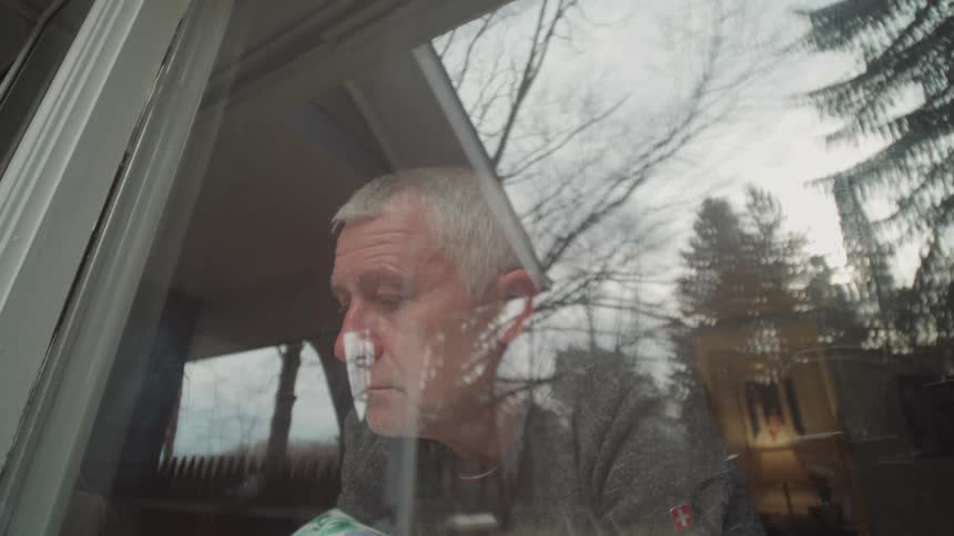 It shows a man with short gray hair and a beard wearing a gray shirt looking out a window his face is reflected in the glass along with the trees and sky outside he appears to be sad or thoughtful