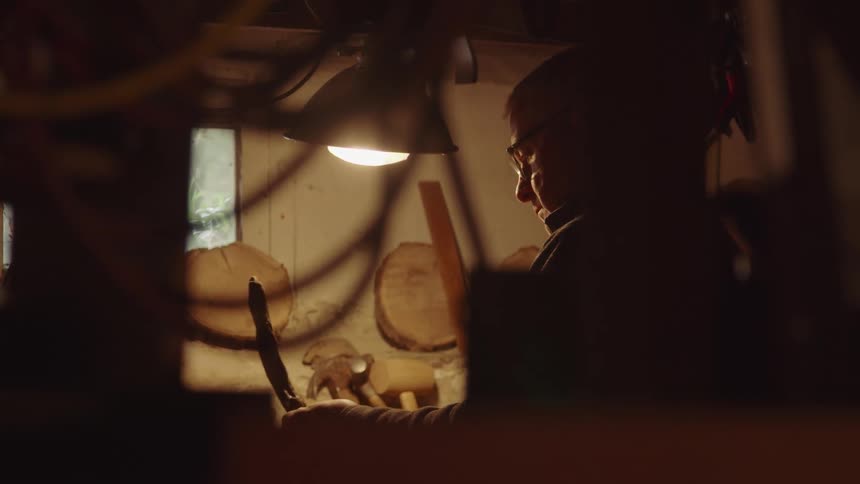 The video shows a man working in a dimly lit workshop illuminated by a single lamp he is focused on a piece of wood carefully shaping it with a tool the warm focused light highlights his concentration on his craft