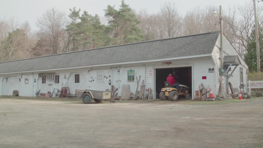 Exterior workshop scene where person rides a lawn tractor at the bay
