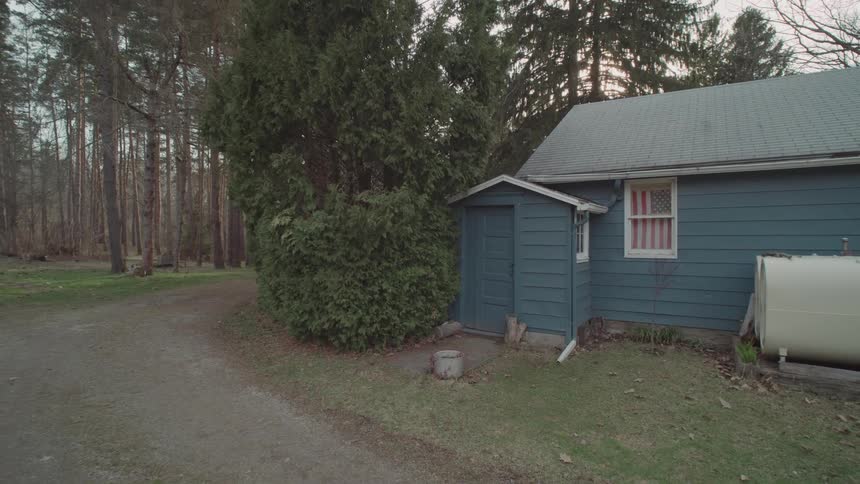 A shot from a drone captures the exterior of a small house with a dark blue roof and siding the house sits on a gravel driveway and is surrounded by a dense forest an american flag is visible in a window on the front of the house