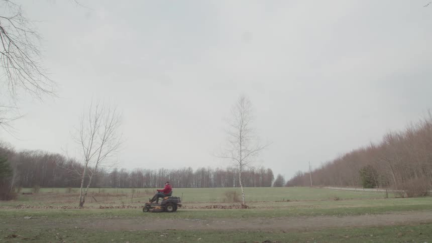 A wide shot captures a person driving a red riding lawn mower across a large field the field is bordered by a line of bare trees on the left and a dense forest on the right under a gray overcast sky