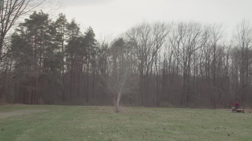 Man on a lawn mower tractor works in a field near a forest