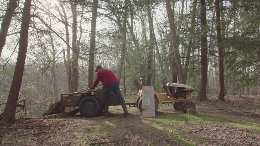 Farmer using a shovel to push dirt from a trailer bed attached to a tractor in the woods at midday