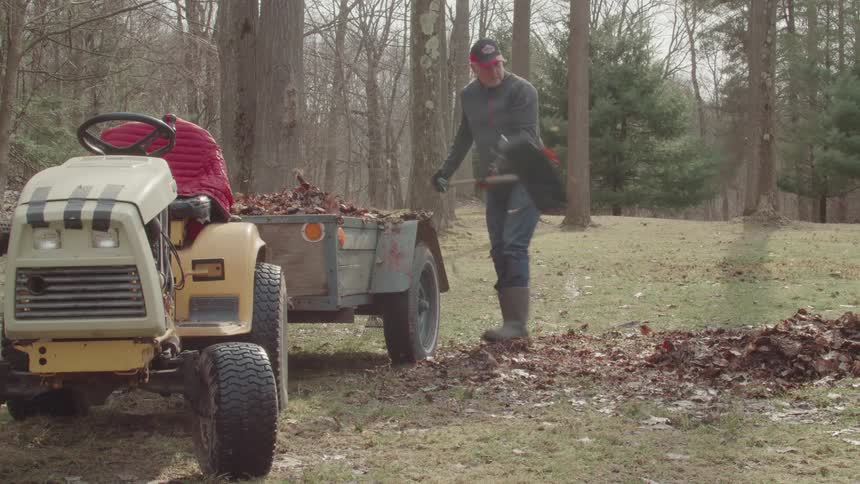 A man is raking and shoveling a large pile of leaves into a trailer attached to a lawn tractor another person is sitting on the tractor helping to organize the leaves in the trailer