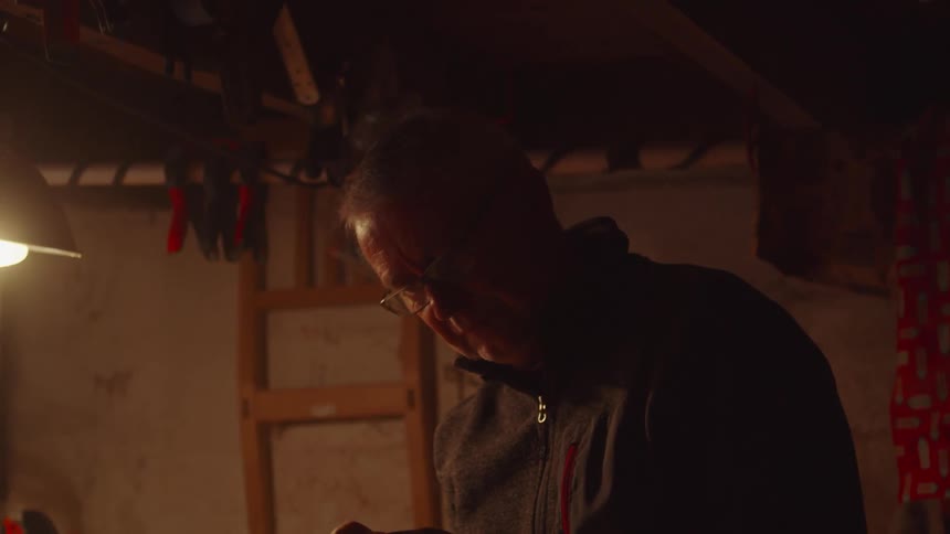 Elderly man wearing glasses and a jacket standing indoors polishing a piece of wood using an electric sander