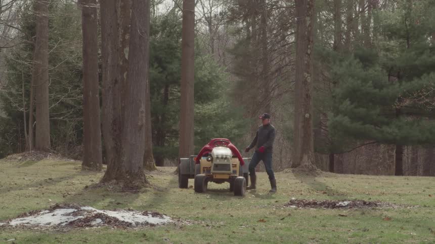 A man shovels leaves in a wooded clearing before getting onto a small yellow tractor