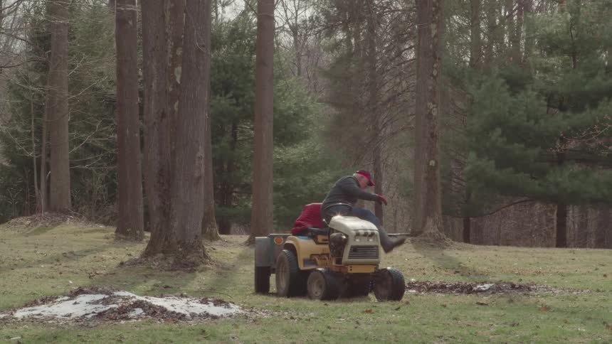 A full shot of a man in a red jacket and a black hat on a lawnmower with a trailer which he drives across a patch of grass in the background are several tall bare trees the man stops the mower gets off and begins to rake leaves into the trailer