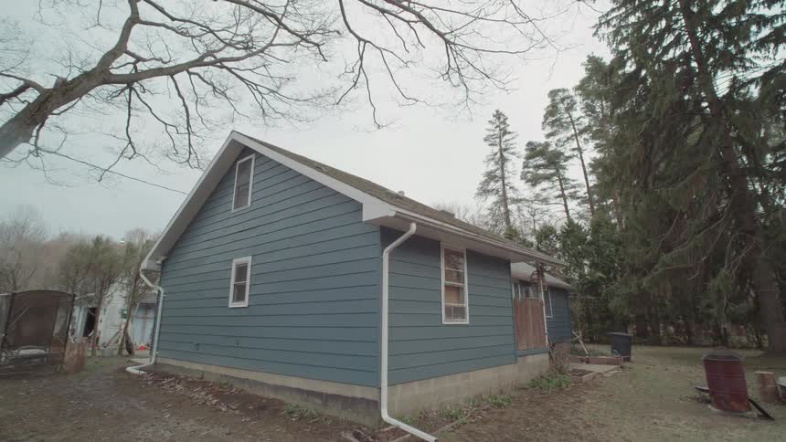 A blue house is shown from the side it has a triangular window on the upper part of the wall the house appears to be located in a rural or suburban area