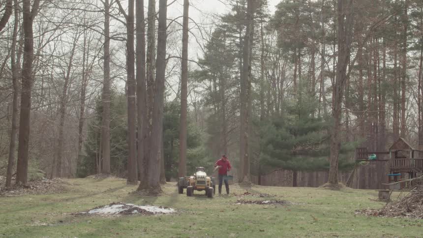 A man in a red jacket and blue jeans is using a rake to pile up leaves in a yard behind him is a large yellow riding mower and a forest of tall mostly bare trees