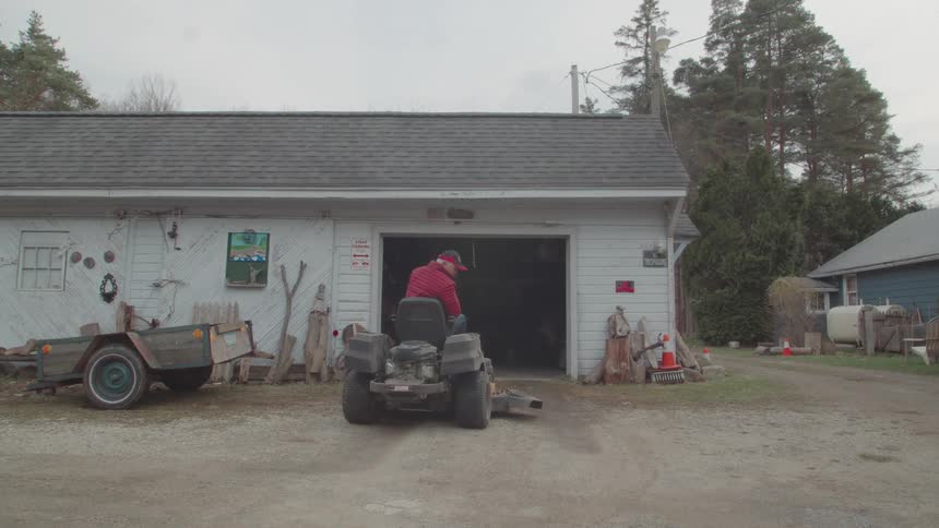 A man wearing a red jacket and hat gets on a riding lawn mower he then backs the mower out of a garage the garage has a large open door and a smaller trailer is parked to the left of the entrance