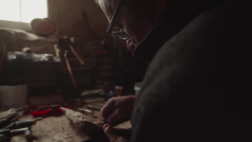 A shot from behind and to the side shows a person wearing glasses and a black jacket leaning over a workbench they are using small tools to carefully carve a piece of wood the workshop is dimly lit with a single window casting a warm light