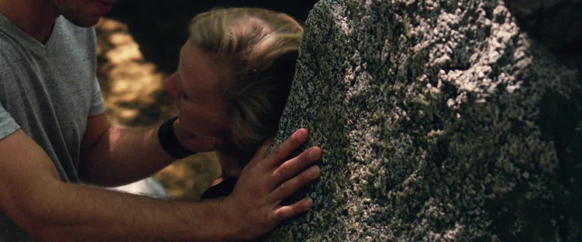 A woman leans against a large rock while a man touches her face