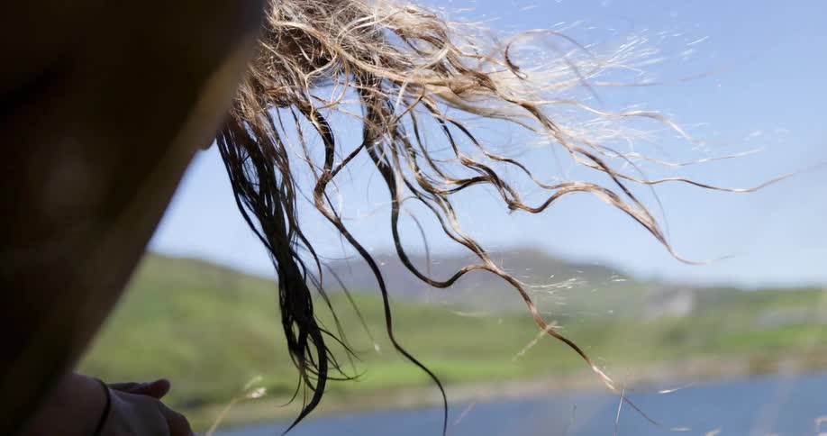 Man with long blond hair leaning on grass beside a lake at midday wind blowing through his hair