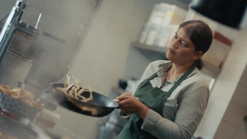 Woman wearing a green apron cooks by tossing food in a frying pan