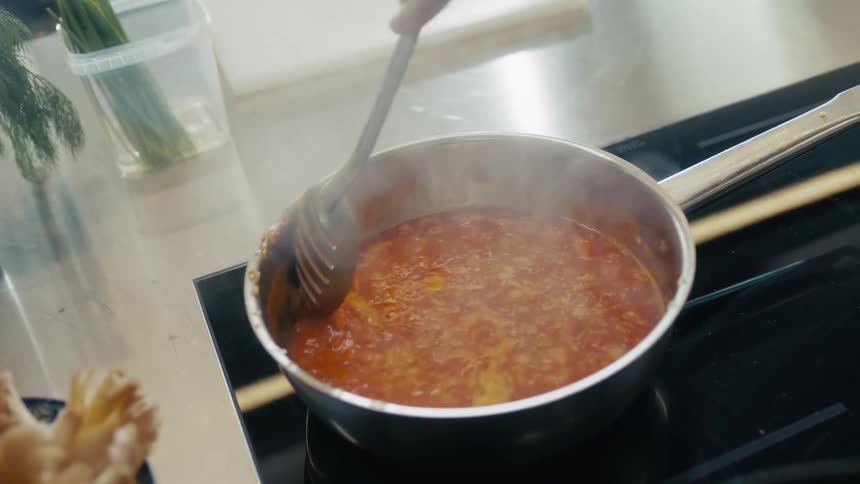 A close view of a persons hand stirring a simmering red sauce with a black slotted spoon in a silver saucepan on a stove