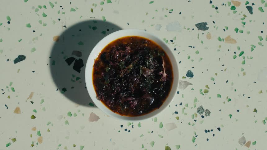 A topdown shot shows a persons hand holding a white bowl filled with a dark reddishbrown dish the bowl is placed on a speckled lightcolored countertop the person sets the bowl down and removes their hand leaving the dish in the center of the fram