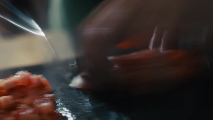 The video shows a closeup of a person using a large knife to chop bright red tomatoes on a dark cutting board the knife is moved with quick efficient strokes