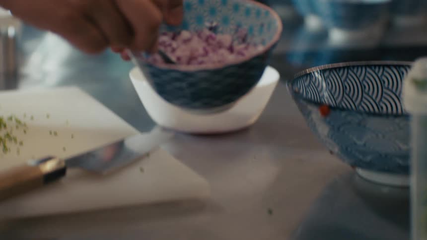 Hand holds a bowl with chopped red onion over a white bowl with tomatoes