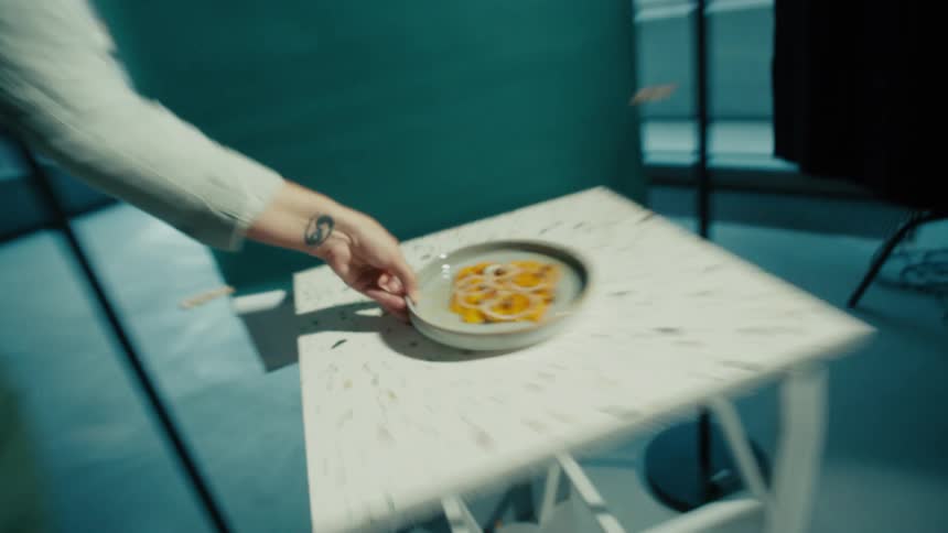 The video shows a person carrying a white dish with food on it they place the dish which contains what appears to be sliced onions in a sauce onto a small white table with a speckled pattern