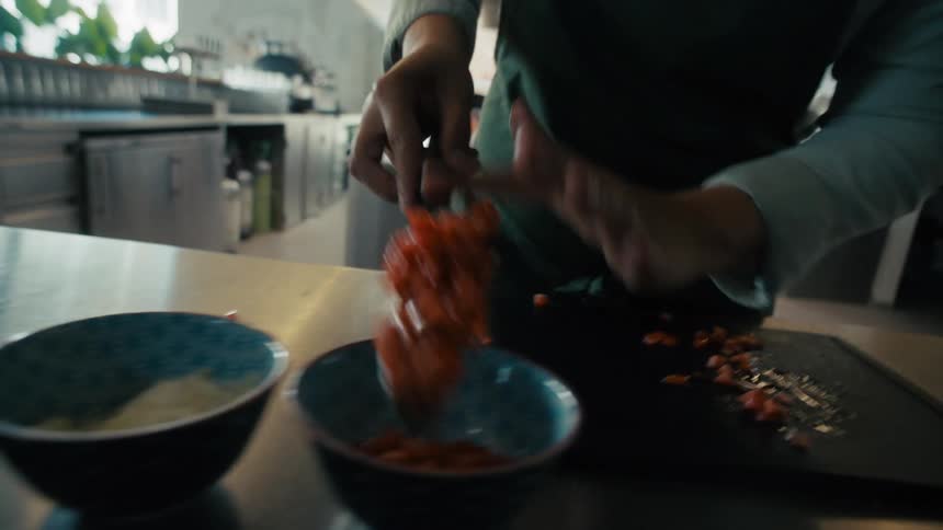 Person transfers chopped tomatoes from the cutting board to a bowl in a kitchen