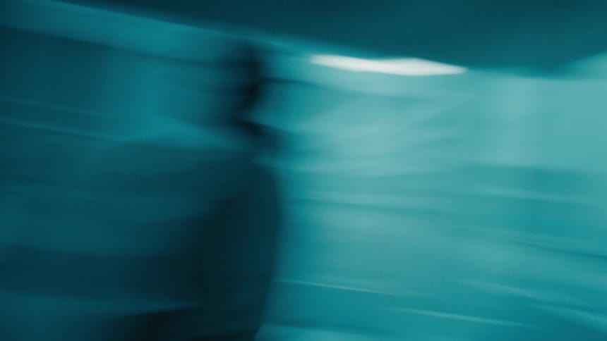 The video shows the silhouette of a person walking along a window with blue light and blurry reflections the persons movement is slightly out of focus creating a sense of being in motion