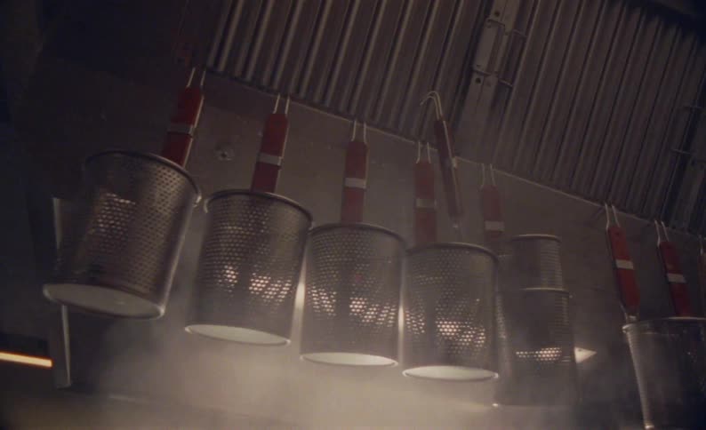 A row of metal pasta strainers hangs from a rack with steam rising from a source below in a dark moodylit commercial kitchen
