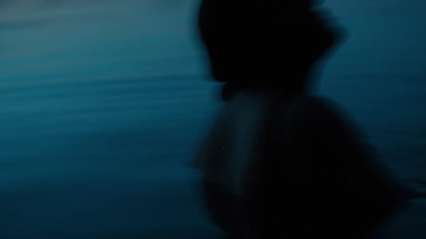 The video shows the silhouette of a person wading into a dark lake at dusk the water is a dark blue almost black color with ripples appearing around them the persons form is mostly obscured by the low light