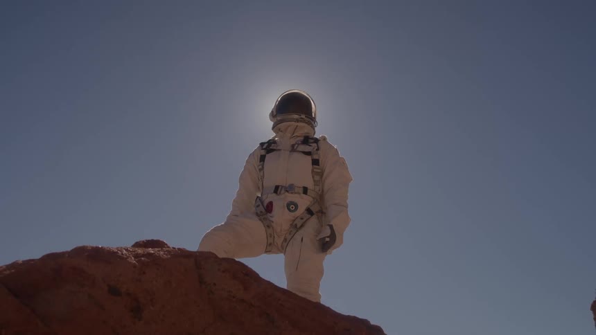A person in a white astronaut suit with a helmet stands on a rocky hill in a desolate landscape with the sun brightly shining behind them