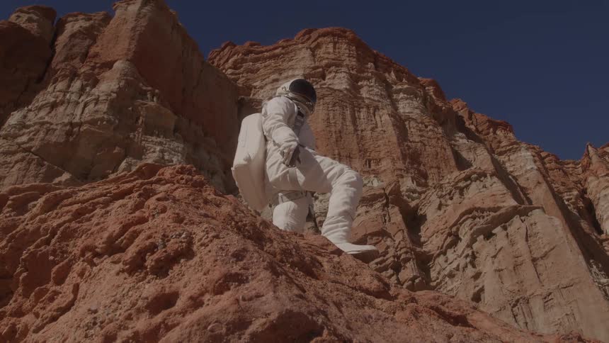 A person in a space suit stands on rocks with canyons in the background